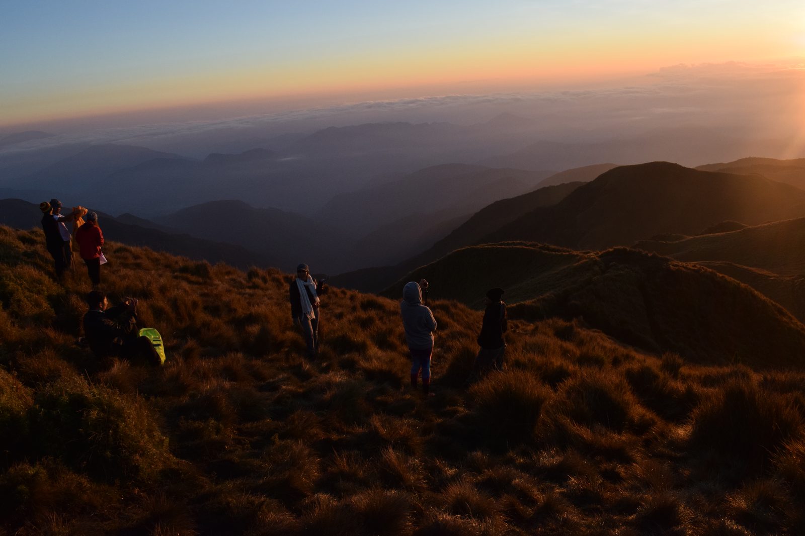 Sekelompok pendaki duduk dan berdiri di puncak gunung menyaksikan matahari terbit di atas lautan awan dan punggungan pegunungan